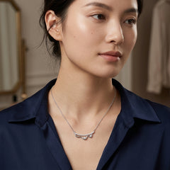 A close-up of a woman wearing a delicate silver necklace with a small bow-shaped pendant set with clear stones, paired with a dark navy blue collared shirt.