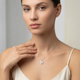 A close-up of a woman wearing a delicate silver necklace with a small round pendant featuring a white floral or starburst design, styled with a cream camisole against a soft, light gray background.