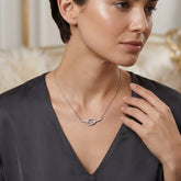 A close-up lifestyle shot of a woman wearing a delicate silver necklace with a central oval link paved with small clear stones, paired with a dark gray V-neck top.
