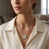 A close-up of a woman wearing a delicate silver four-leaf clover pendant necklace over a cream-colored collared blouse, with a softly blurred indoor background.