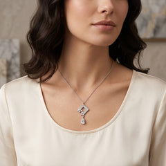 A close-up of a woman wearing a delicate silver pendant necklace with a geometric diamond design and a teardrop crystal drop, paired with a cream-colored satin top.