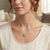 A close-up of a woman wearing a delicate silver heart pendant necklace with a clear stone, styled with a cream-colored scoop-neck top.