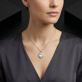 A close-up of a woman wearing a silver-toned necklace with a circular pendant featuring a heart motif, paired with a dark gray satin top.