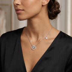 A close-up lifestyle shot of a woman wearing a silver necklace with a double-ring pendant and a small disc charm, paired with a black satin V-neck top.