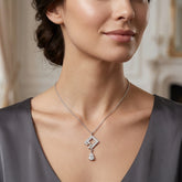 A close-up of a woman wearing a delicate silver necklace with a geometric diamond pendant and a teardrop crystal drop, set against a softly blurred indoor background.