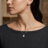 A close-up lifestyle shot of a woman wearing a delicate silver chain necklace with a small, round, clear gemstone pendant. The pendant features an engraved cross design.