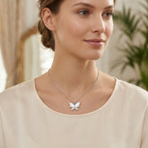 A woman wearing a delicate silver butterfly pendant necklace, styled with a cream-colored top. The background is a softly blurred indoor setting with warm neutral tones and a hint of green foliage.