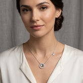 A woman wearing a delicate silver necklace with a circular pendant featuring a central dark stone and a small round charm, paired with a white blouse against a soft gray background.