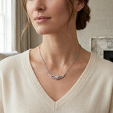 A close-up of a woman wearing a delicate silver necklace with a geometric curved pendant featuring a central clear rectangular stone, paired with a cream-colored V-neck sweater.