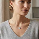 A woman wearing a delicate silver chain necklace with a small, sparkling heart-shaped pendant, styled with a light gray V-neck sweater in a softly lit indoor setting.