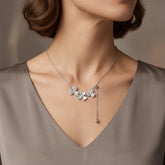 A close-up of a woman's neck and upper chest wearing a delicate silver necklace with a cluster of small flower-shaped charms and a dangling bead, paired with a taupe V-neck top.