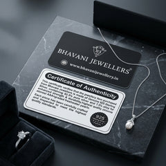A silver pearl necklace with a delicate chain and bezel-set pearl pendant is displayed on a dark marble surface next to its black presentation box, a black Bhavani Jewellers brand card, and a Certificate of Authenticity. A velvet ring box with a silver ring is partially visible in the lower-left corner.