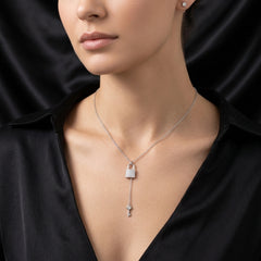 A close-up of a model wearing a silver key-shaped pendant necklace with a padlock at the top, set against a black satin background.