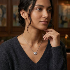 A woman wearing a delicate silver heart pendant necklace with a clear stone, styled with a dark gray V-neck sweater in a warm, softly blurred indoor setting.