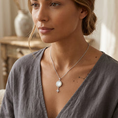 A woman wearing a delicate silver chain necklace with a white circular pendant and a small star charm, styled with a gray linen top in a softly lit indoor setting.