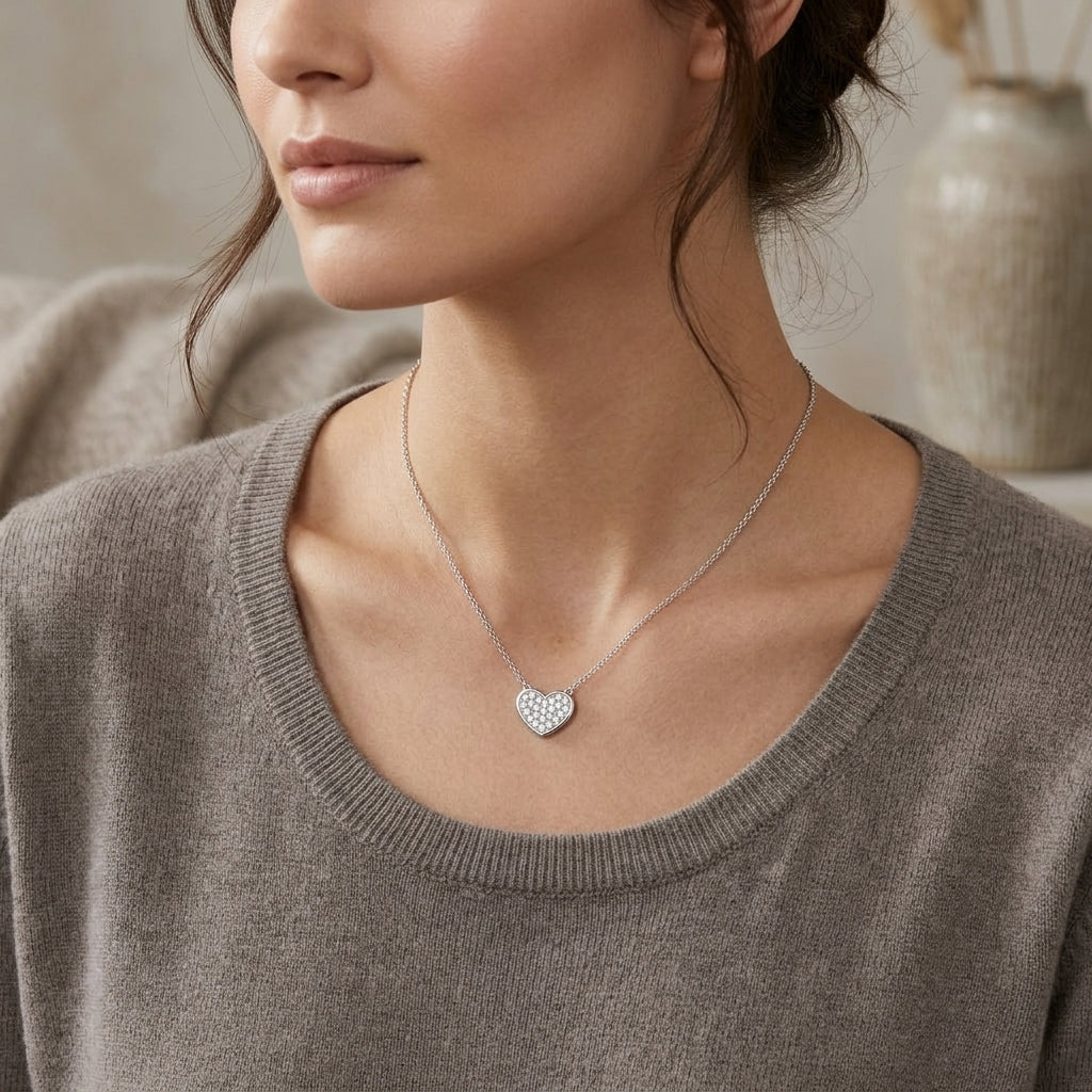 A close-up lifestyle shot of a woman wearing a delicate silver heart pendant necklace with a pavé-set design, styled with a taupe knit sweater.