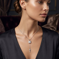 A woman wearing a silver-toned Y-necklace with a padlock and key pendant, shown in a close-up portrait against a dark, elegant interior background.