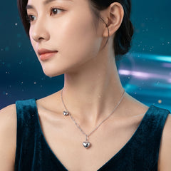A woman wearing a delicate silver heart pendant necklace with a small matching heart charm, shown in a close-up portrait against a dark blue, starry background.