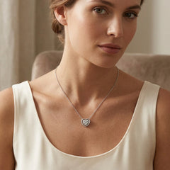 A woman wearing a delicate silver heart pendant necklace with a pavé-set center, styled with a cream-colored satin top. The background is a softly blurred indoor setting with neutral beige tones.