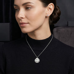A woman wearing a delicate silver pendant necklace with a circular cutout design and a matching stud earring, dressed in a black mock-neck top against a dark background.