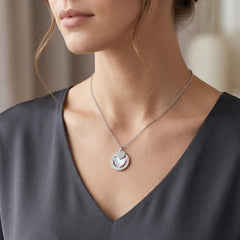 A close-up of a woman wearing a delicate silver necklace with a circular pendant featuring a small heart and a textured outer ring, styled with a dark gray V-neck top.