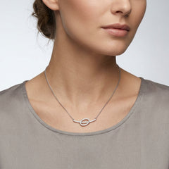 A close-up of a woman wearing a delicate silver chain necklace with a small, oval, diamond-set pendant. She is wearing a taupe-gray top against a clean, light gray background.