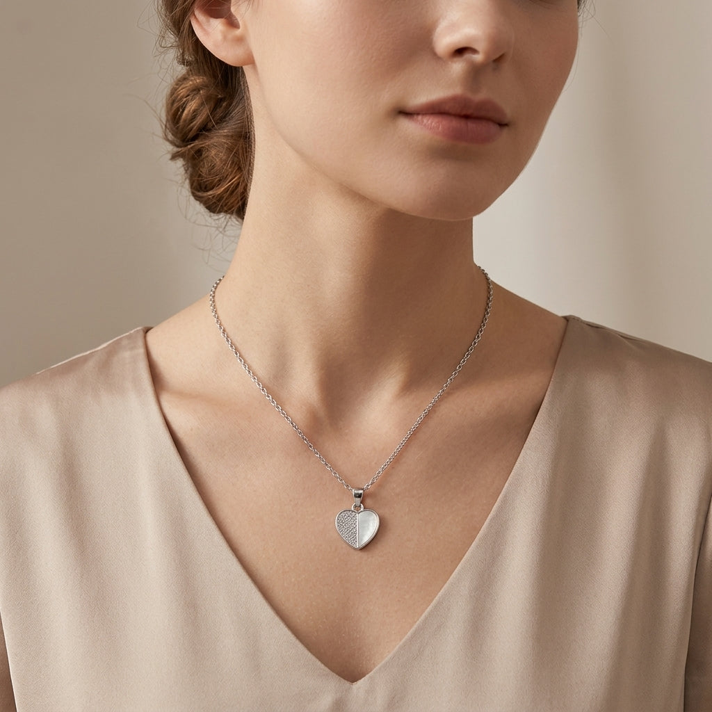 A close-up of a woman wearing a delicate silver chain necklace with a textured heart-shaped pendant, styled with a beige satin V-neck top.