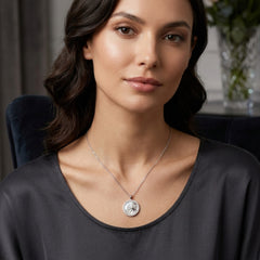 A woman wearing a delicate silver chain necklace with a round silver pendant featuring a sun-like emblem, photographed in a softly lit indoor setting.