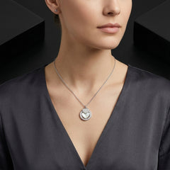 A close-up of a woman wearing a silver-toned necklace with a circular pendant featuring a heart motif, paired with a dark gray satin top.