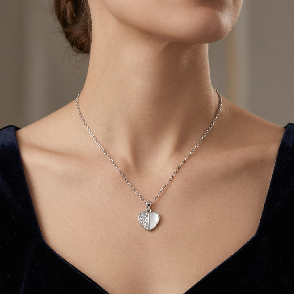 A close-up of a model wearing a delicate silver heart pendant necklace. The necklace features a fine chain and a small, polished heart-shaped pendant with a textured pattern on one side.