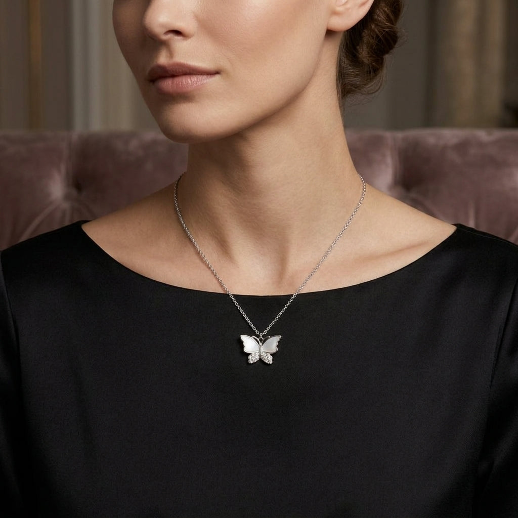 A close-up of a woman wearing a delicate silver butterfly pendant necklace on a fine chain, paired with a black satin top and a soft mauve tufted chair in the background.