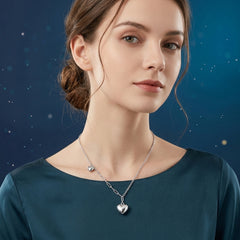 A woman wearing a silver heart pendant necklace with a delicate chain and a small matching heart charm, set against a dark blue starry background.