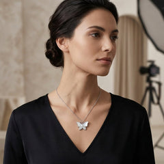 A woman wearing a delicate silver butterfly pendant necklace, styled with a black V-neck top. The background is a softly blurred studio setting with warm beige tones and a softbox light.