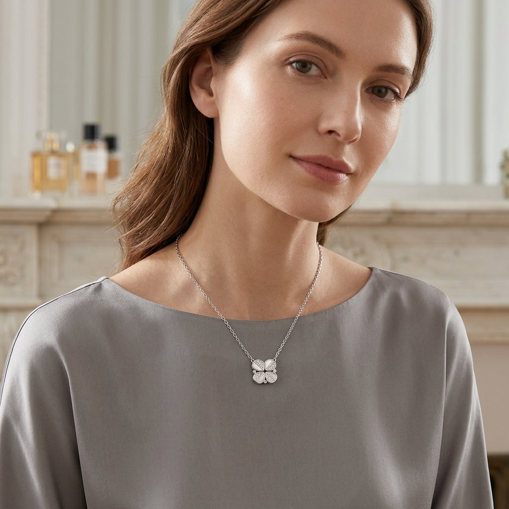 A woman wearing a delicate silver necklace with a four-petal flower pendant, styled with a gray satin top in a softly lit indoor setting.