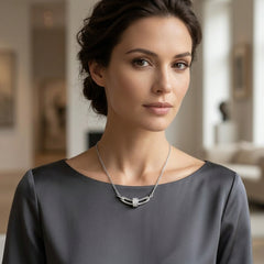A woman with dark hair in a low bun wearing a silver-toned geometric chain necklace with a rectangular center, paired with a dark gray satin blouse. The background is a softly blurred, warm-toned interior room.