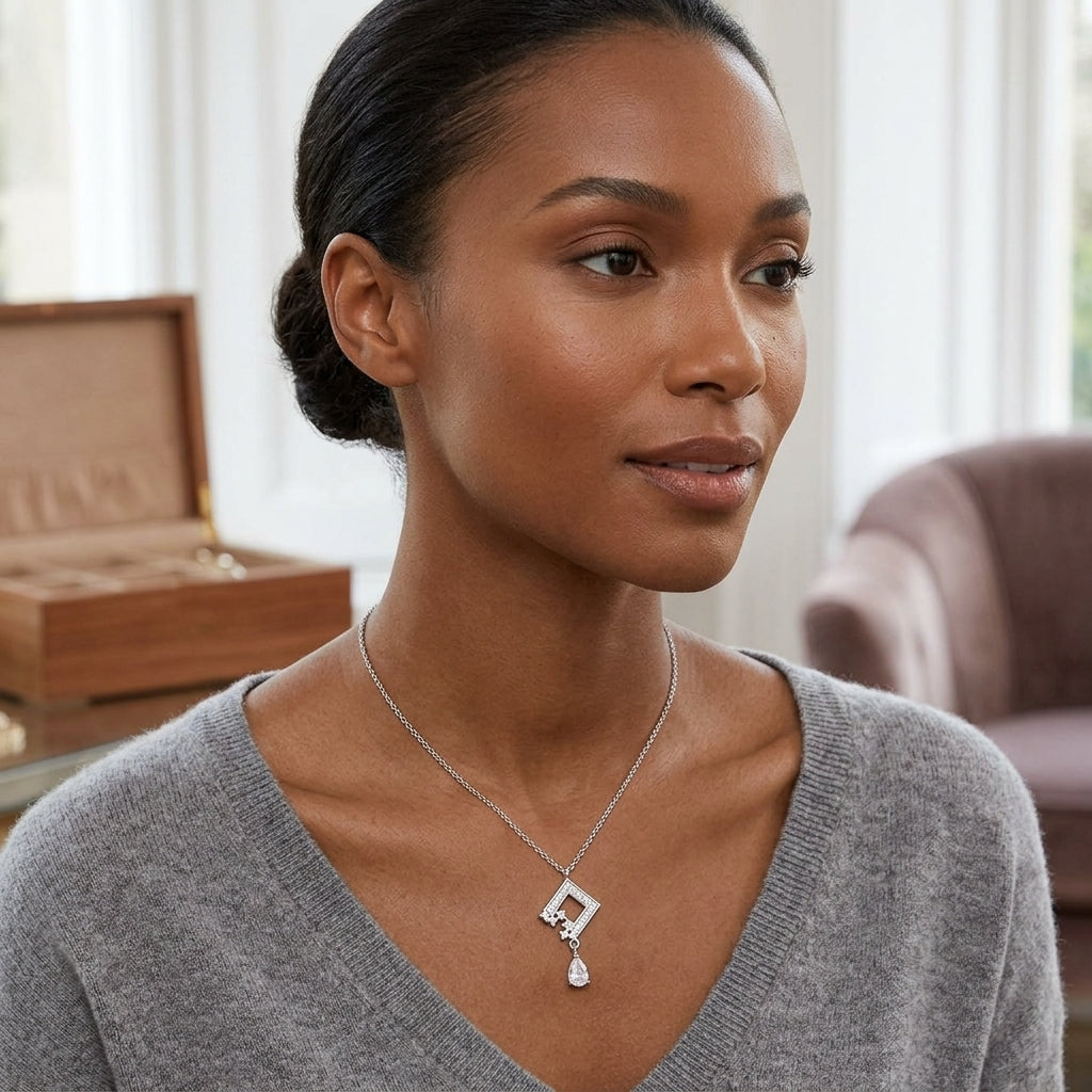 A woman wearing a delicate silver geometric pendant necklace with a teardrop charm, styled with a gray V-neck sweater in a softly lit indoor setting.