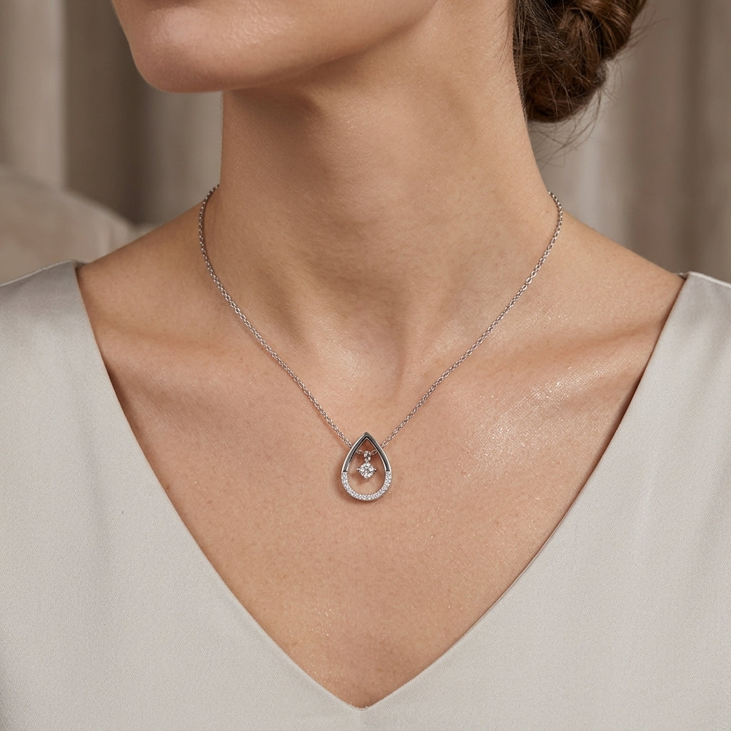 A close-up of a woman wearing a delicate silver teardrop pendant necklace with a small central pearl, styled with a light beige V-neck top.