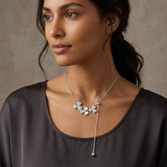 A woman wearing a delicate silver lariat necklace with a cluster of small white flower charms and a dangling bead at the end, paired with a dark gray top against a neutral textured background.