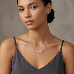 A woman wearing a delicate silver necklace with a curved, wing-like pendant, styled with a dark gray camisole against a neutral gray background.
