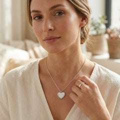 A woman wearing a delicate silver heart pendant necklace with a textured, pave-like surface detail, styled with a matching silver ring on her finger. She is wearing a soft cream-colored top in a warm, softly lit indoor setting.