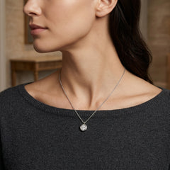 A close-up lifestyle shot of a woman wearing a delicate silver chain necklace with a small, round, clear gemstone pendant. The pendant features an engraved cross design.