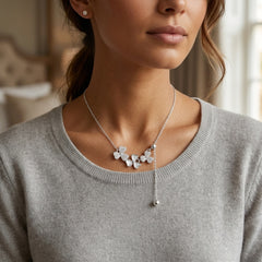 A close-up of a woman wearing a delicate silver necklace with three small flower charms and a dangling bead, paired with a gray knit top.