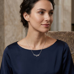 A woman wearing a delicate silver necklace with a small curved bar pendant, paired with a navy blue top, shown in a softly lit indoor setting.