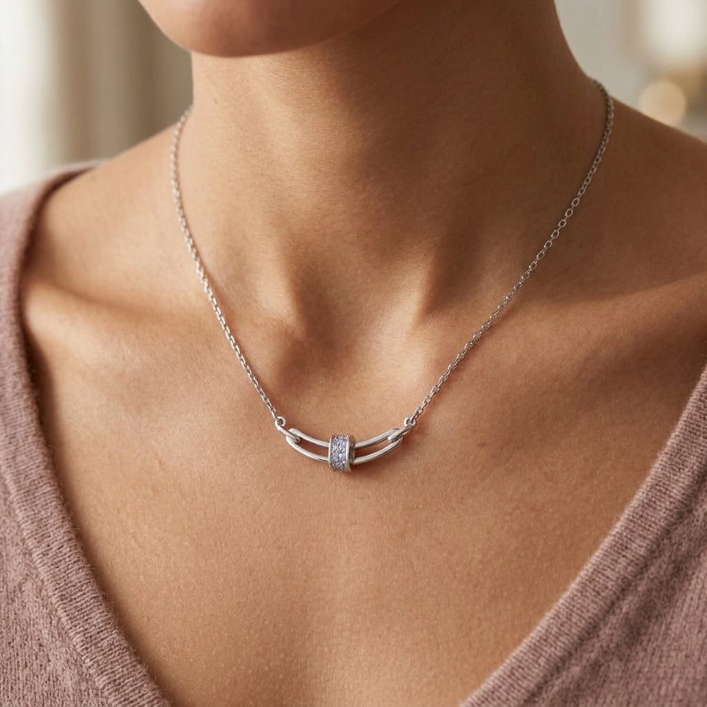 A close-up of a model wearing a delicate silver necklace with a curved double-bar pendant featuring a small clear stone cluster in the center, styled with a mauve knit top.