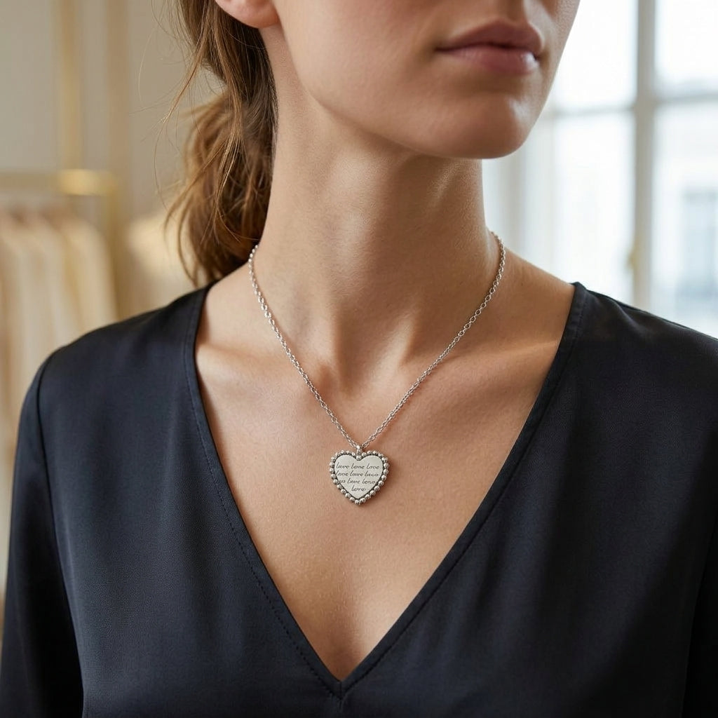 A close-up of a woman wearing a silver heart pendant necklace with a textured pattern, styled with a black satin V-neck top.