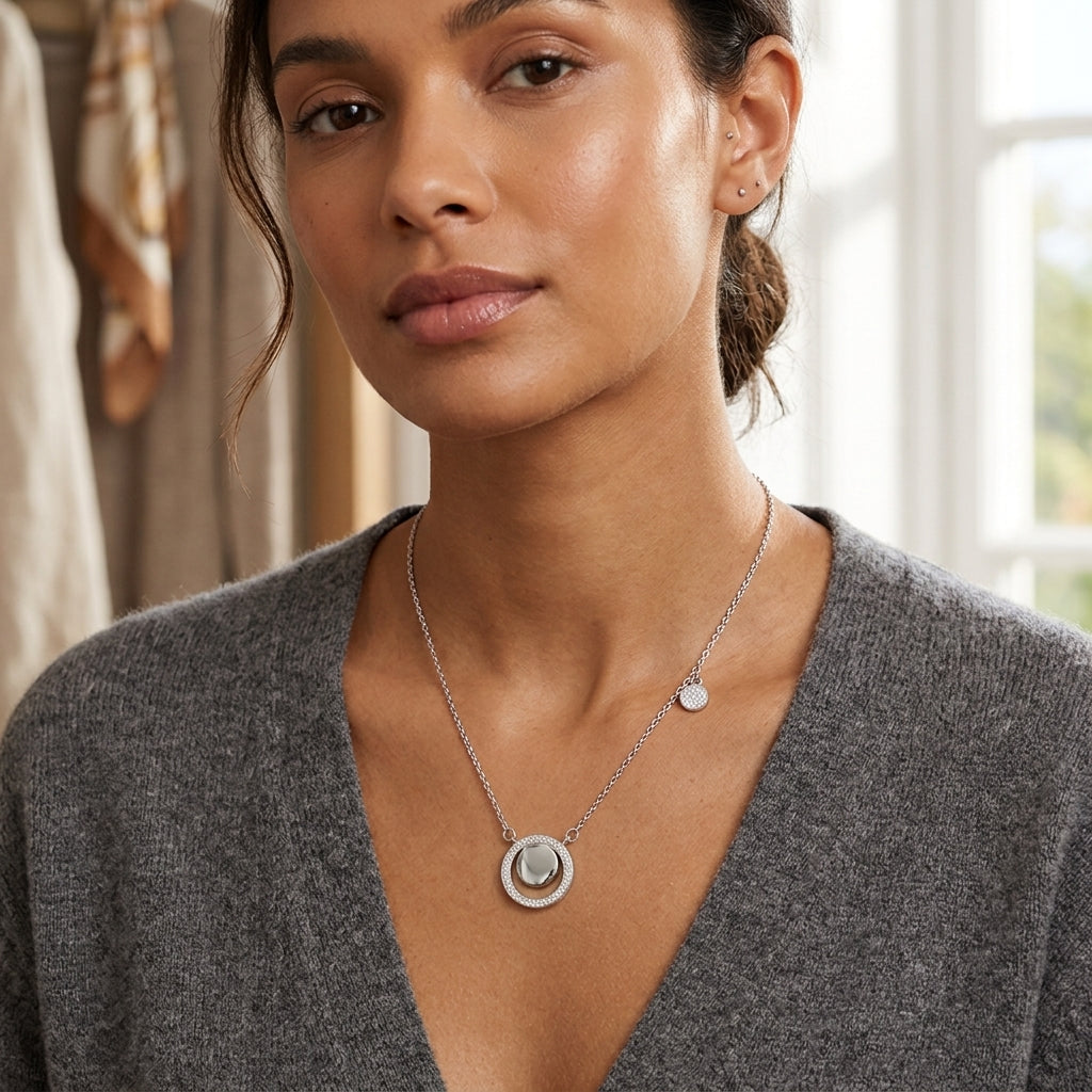 A woman wearing a silver necklace with a double-ring pendant and a small disc charm, styled with a gray V-neck sweater in a softly lit indoor setting.