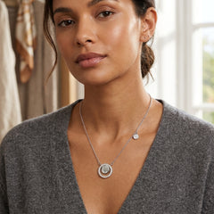 A woman wearing a silver necklace with a double-ring pendant and a small disc charm, styled with a gray V-neck sweater in a softly lit indoor setting.