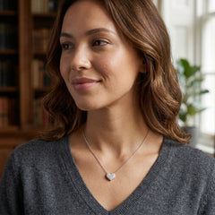 A woman with wavy brown hair wearing a delicate silver heart pendant necklace, shown in a warm indoor setting with a blurred bookshelf in the background.