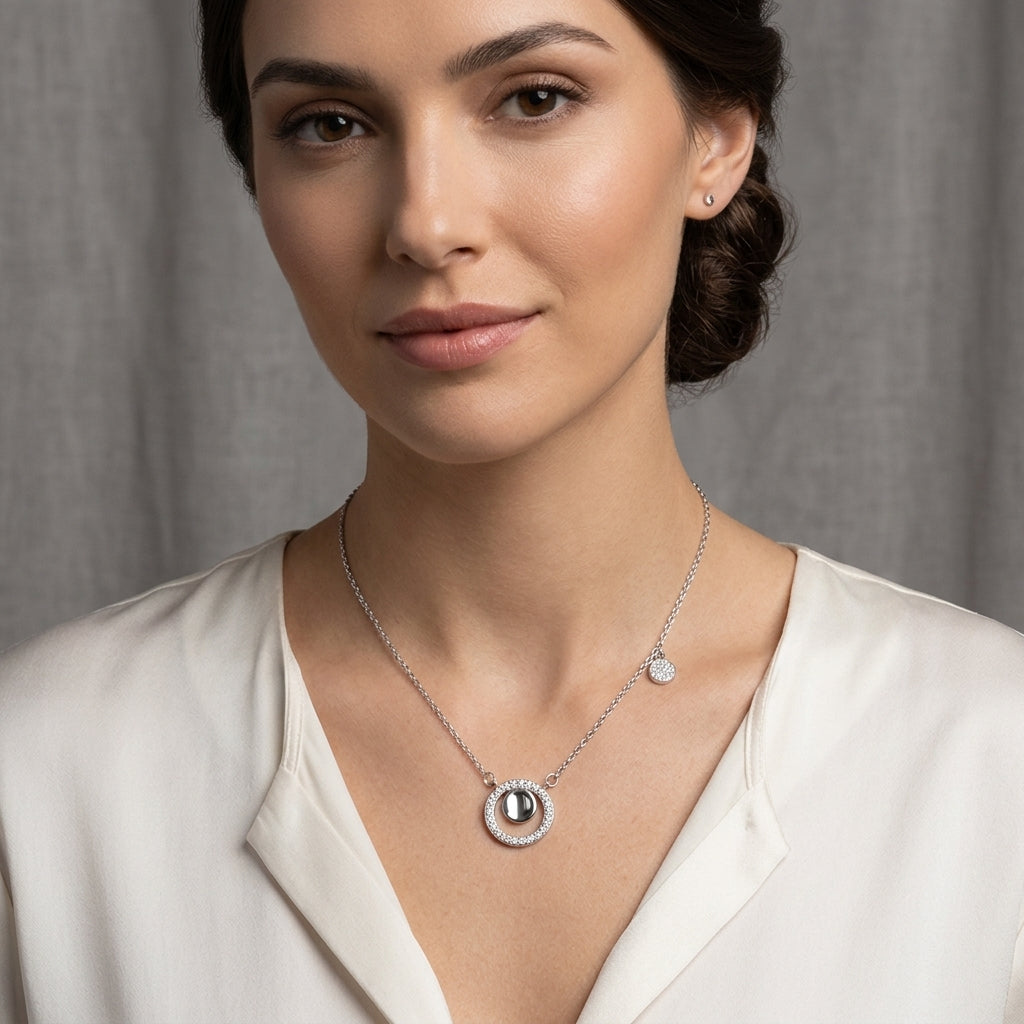 A woman wearing a delicate silver necklace with a circular pendant featuring a central dark stone and a small round charm, paired with a white blouse against a soft gray background.
