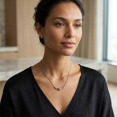 A woman wearing a delicate silver necklace with a modern geometric bar pendant, styled with a black V-neck top in a softly lit indoor setting.